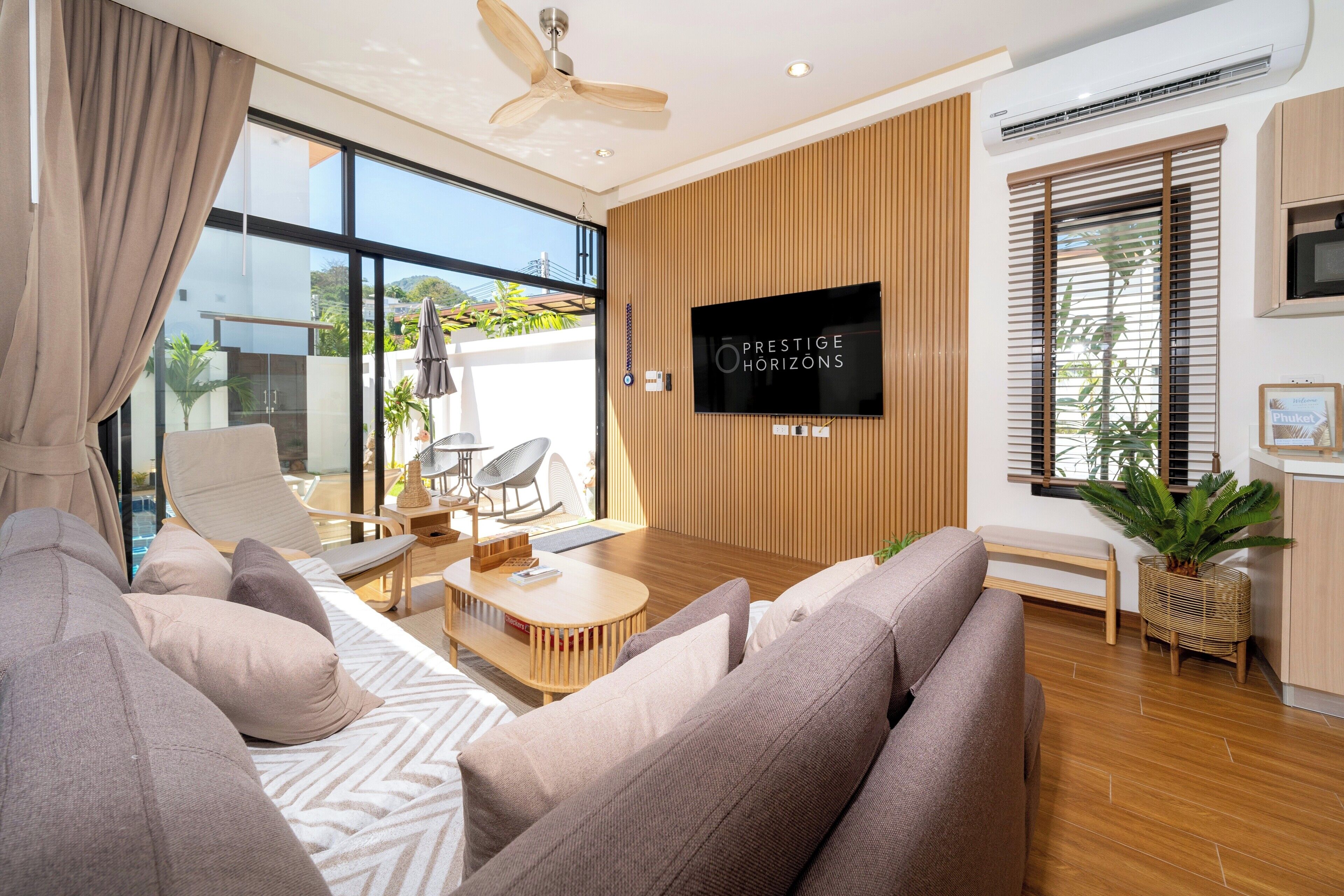 A modern living room with a sofa, coffee table, and TV. There is a large window with blinds, a ceiling fan, and a plant.
