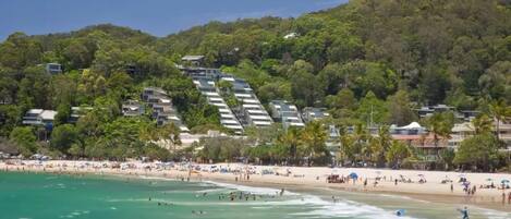 A beach with a hillside of buildings, clear turquoise water, and a crowd of people.