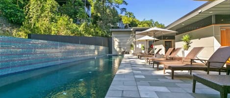 Piscina com espreguiçadeiras e guarda-sóis, rodeada por vegetação e um edifício moderno.