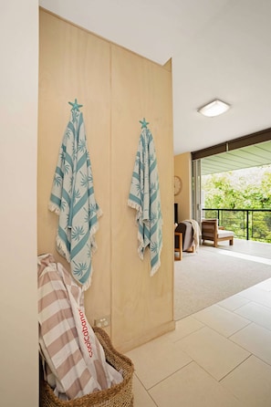 Una habitación con un armario de madera, dos toallas colgadas en ganchos, una cesta de mimbre con un cojín a rayas, y vistas a un balcón con una silla y una mesa.