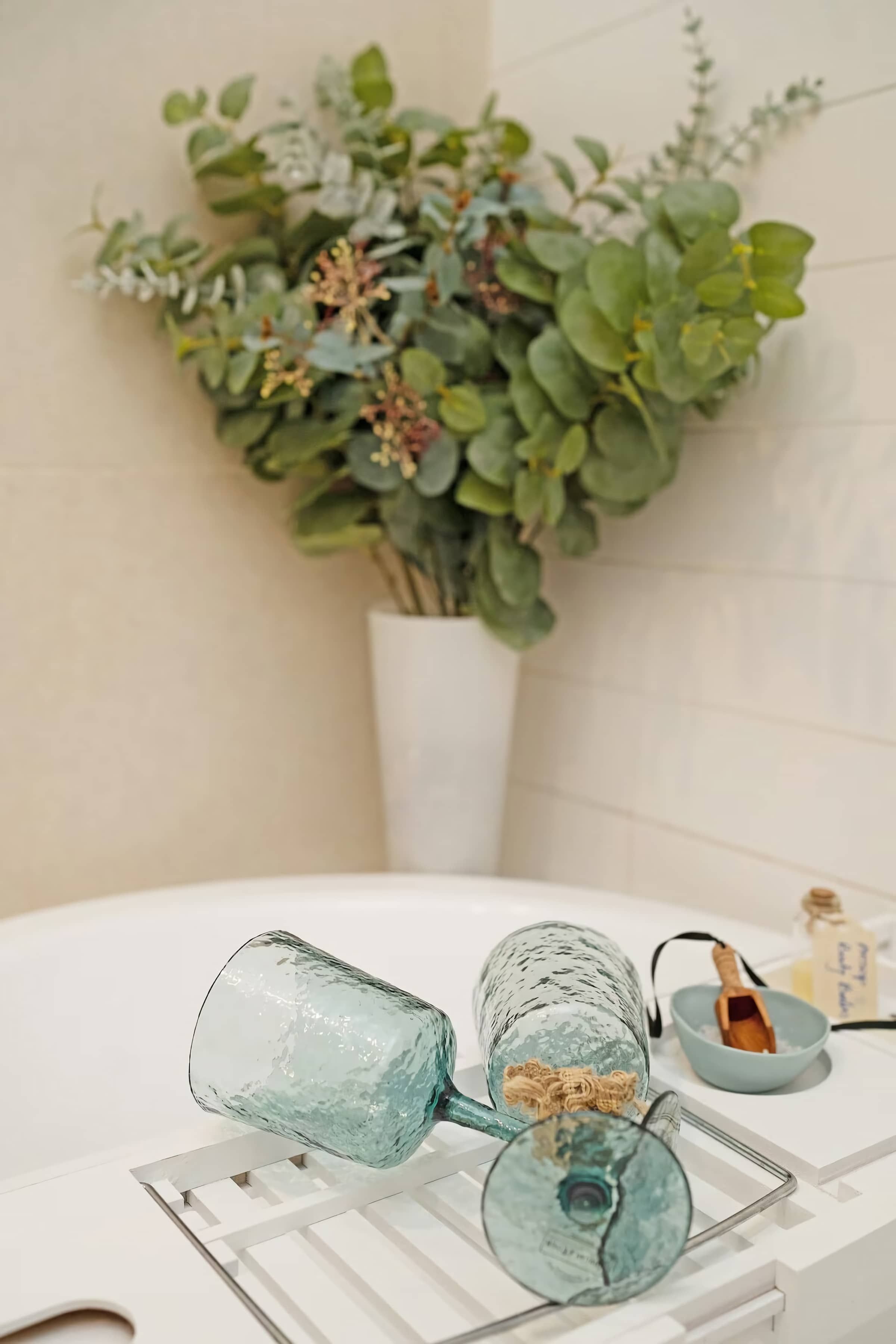 Un baño con un lavamanos blanco, un florero de vidrio con plantas verdes y un cuenco de madera pequeño con una cuchara.