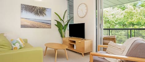 A living room with a sofa, a coffee table, a TV stand, and a balcony with a view of trees.