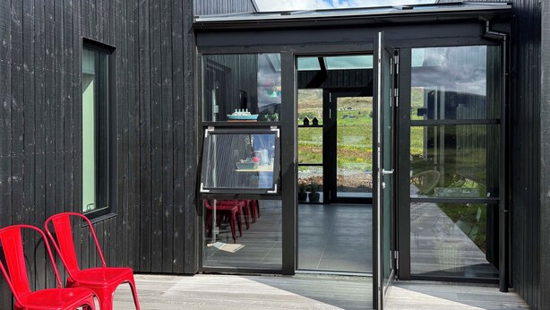 A modern building with a glass door entrance, red chairs outside, and a view of a green landscape. - Entire home in Grímsnes og Grafningshreppur, Iceland (Grímsnes  og Grafningshreppur)