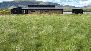 A modern building with a flat roof, large windows, and a wooden exterior, situated in a grassy field with mountains in the background. - Entire home in Grímsnes og Grafningshreppur, Iceland (Grímsnes  og Grafningshreppur)