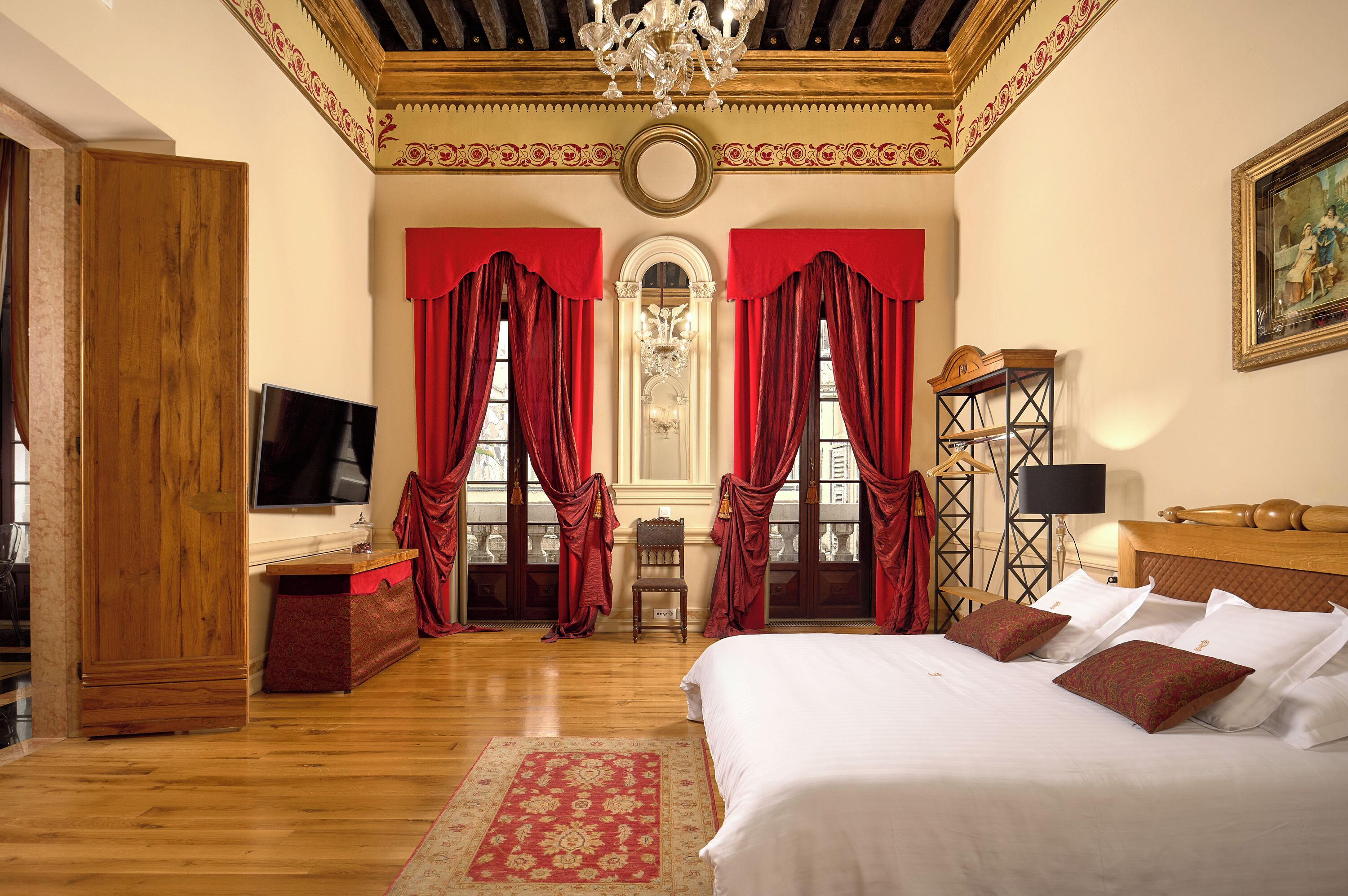 A hotel room with a wooden floor, a large bed with white linens, red curtains, a chandelier, and a painting on the wall.