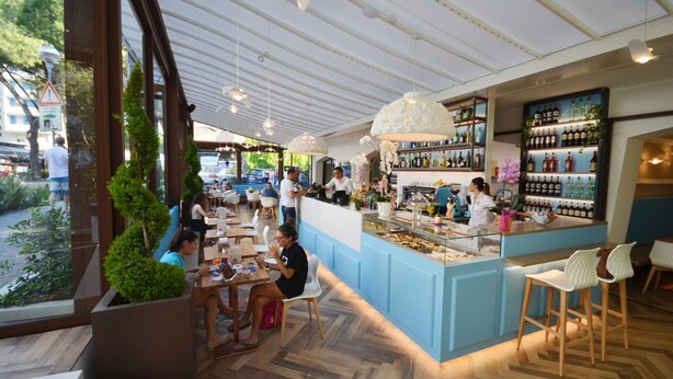 Un café interior con suelo de madera, una zona de bar con vitrina de vidrio y diferentes disposiciones de asientos.