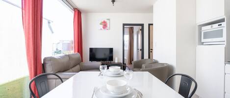 Una sala de estar moderna con una mesa de comedor blanca para dos personas, un sofá y un televisor de pantalla plana.