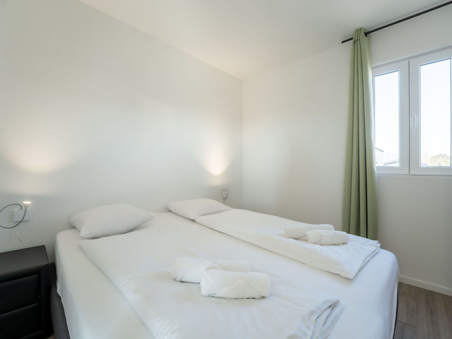A clean, modern hotel room with a large bed, two pillows, a bedside table, and a window with curtains.