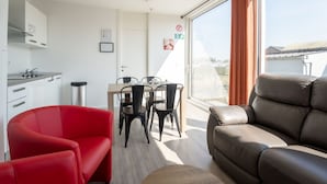 A modern living room with a brown leather sofa, a red chair, and a dining table with four chairs. There is a kitchen area with white cabinets and a stainless steel sink. A large window with orange curtains provides a view of the outside. - Beautiful apartment in villa for 4 guests with WIFI, A/C, TV, terrace and pets allowed (Bruges)