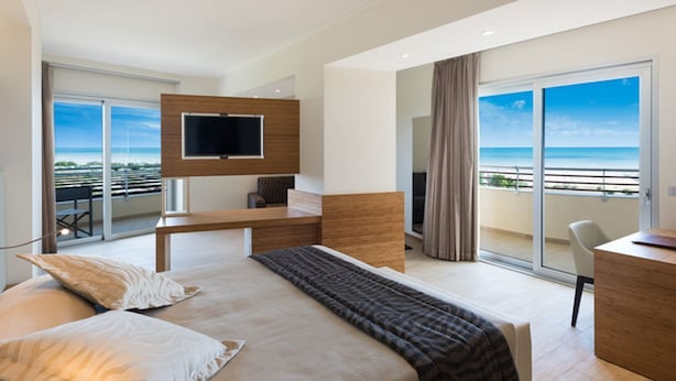 Ein modernes Hotelzimmer mit einem großen Bett, einem Schreibtisch, einem Fernseher und einem Balkon mit Meerblick.