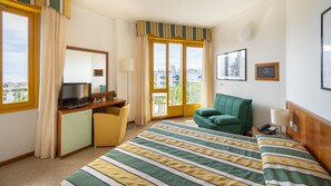 Habitación de hotel con cama, escritorio con televisor, silla, balcón con vistas y ventana con cortinas.