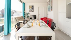 Un comedor moderno con una mesa blanca preparada para el desayuno, un televisor al fondo y vistas al exterior.
