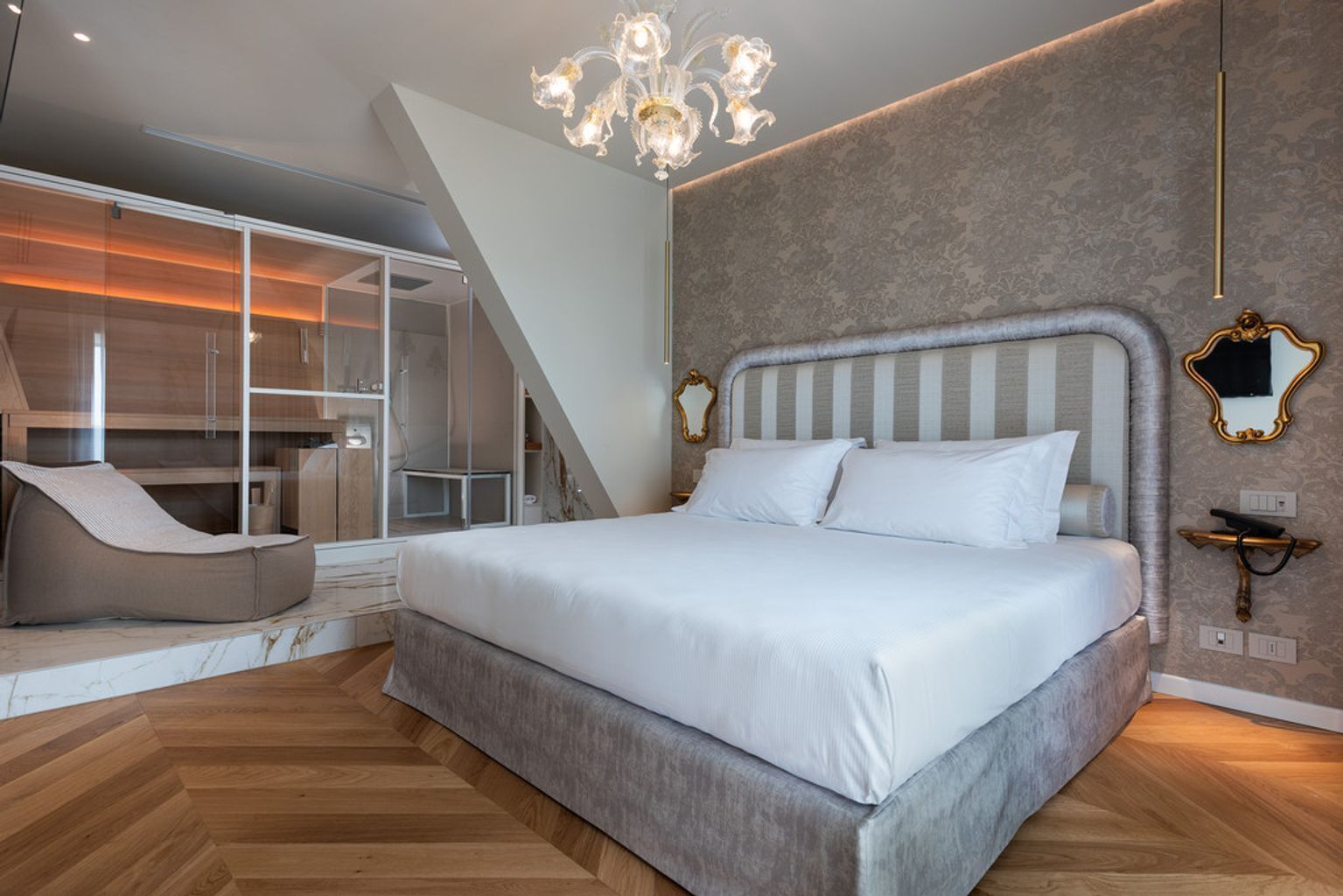 Un dormitorio moderno con una cama grande, un chaise lounge, una ducha con cerramiento de vidrio y una araña.