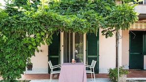 Un patio al aire libre con una mesa y sillas a cuadros, rodeado de abundante follaje y un edificio con contraventanas verdes.