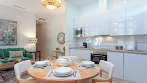 Una cocina moderna con armarios blancos, microondas y pileta. Hay un comedor con una mesa redonda de madera dispuesta para cuatro, sillas blancas y un sofá verde al fondo.