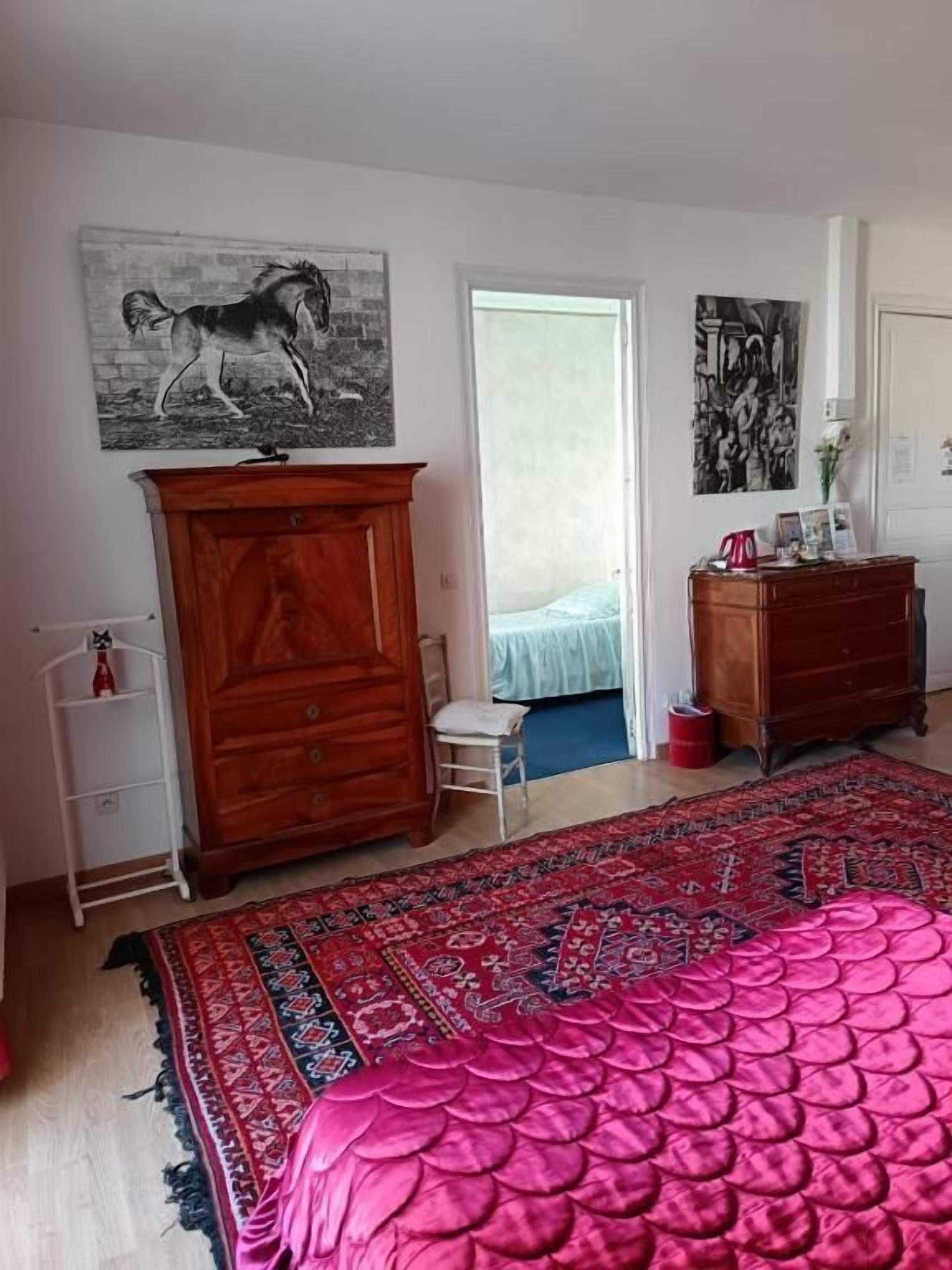 Un dormitorio con una cama grande, un armario de madera, una silla pequeña, un hervidor rojo y una puerta que da a otra habitación.