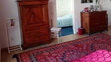 Un dormitorio con una cama grande, un armario de madera, una silla pequeña, un hervidor rojo y una puerta que da a otra habitación.