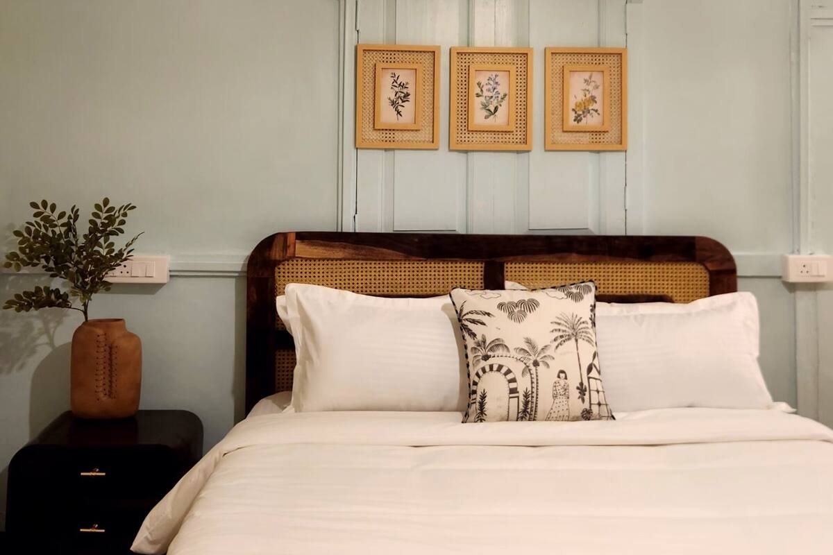 A neatly made bed with white linens and a yellow decorative throw, a wooden headboard, and three framed floral artworks on the wall.