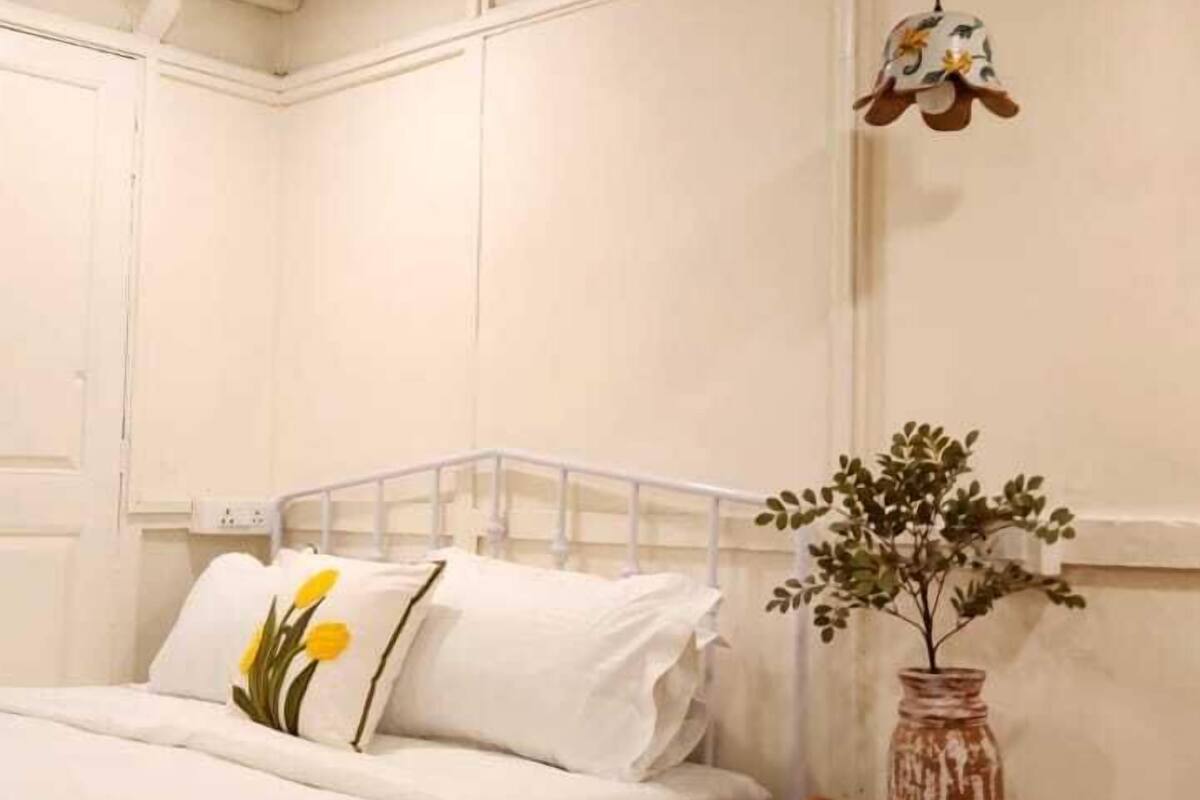 A room with two beds, a small table with a vase, and a wicker chair.