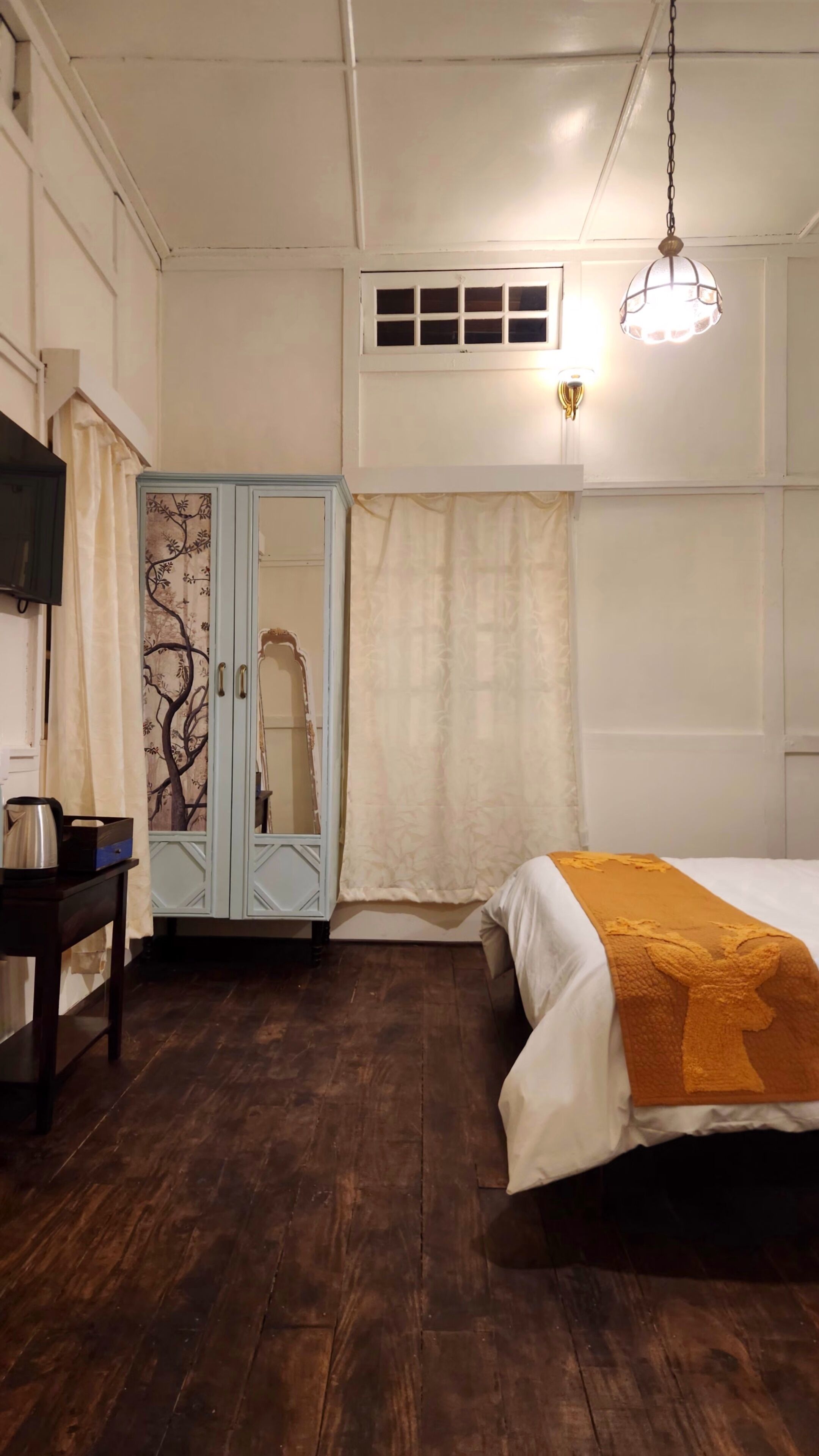 A bedroom with a bed, a wardrobe, a desk, and a hanging light fixture.