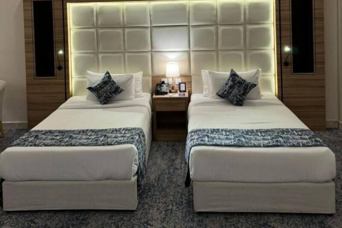 A hotel room with two beds, a large padded headboard, a nightstand with a lamp, and a small table.