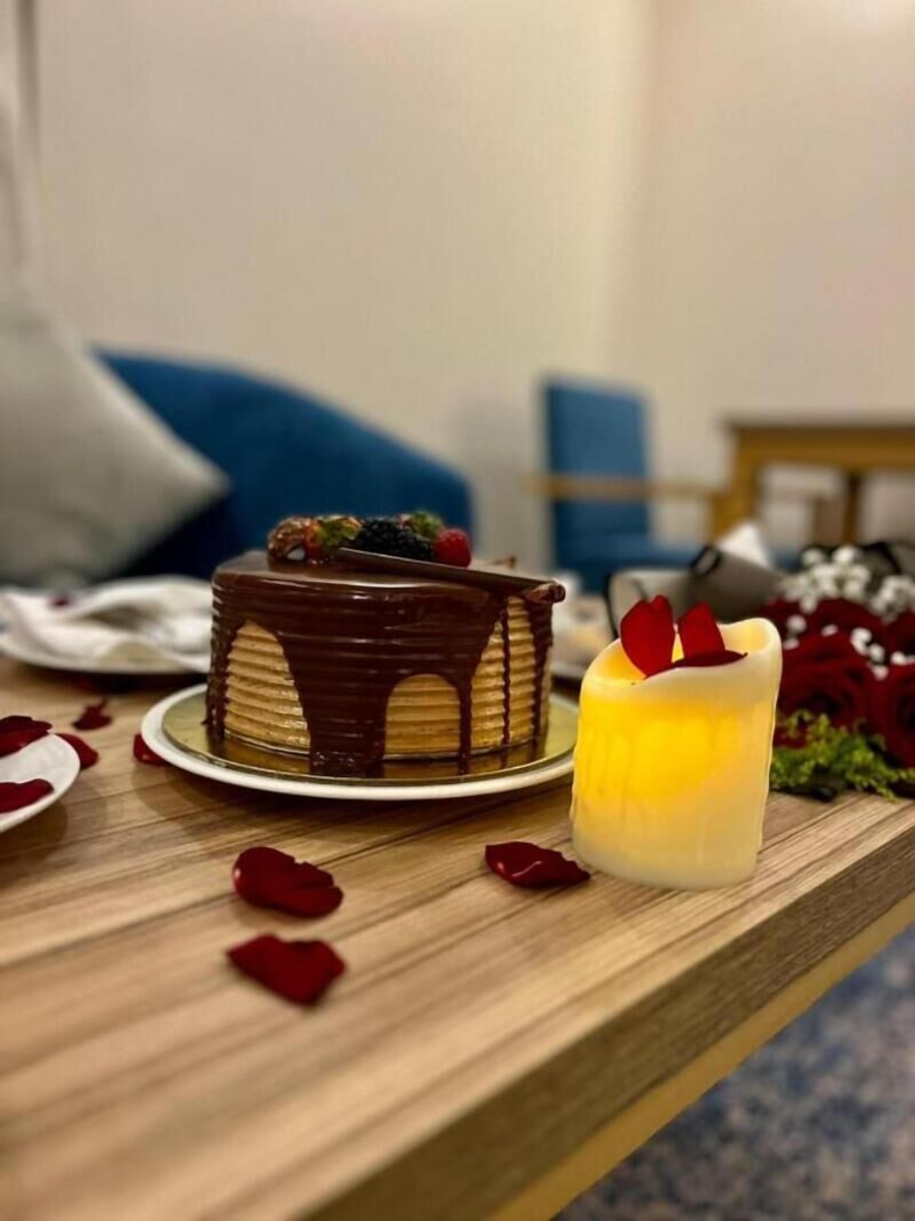 A cake with chocolate drips on a plate, a candle, and rose petals on a wooden table.