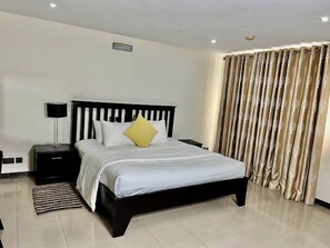 Quarto moderno com cabeceira preta, roupa de cama branca, travesseiro amarelo decorativo, mesa de cabeceira com abajur e janela com cortinas bege.