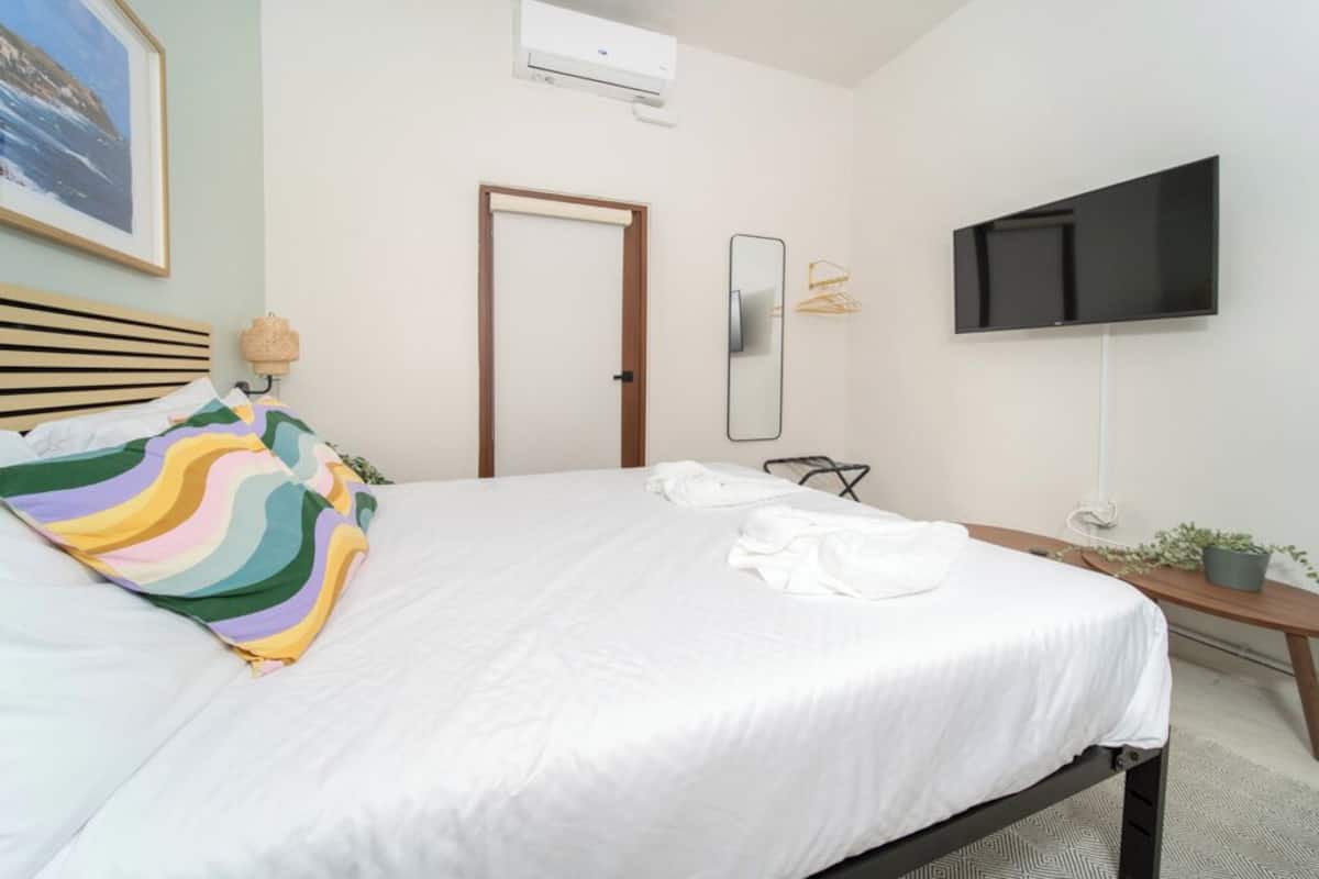 A neatly made bed with white bedding, a wooden headboard, and a colorful striped pillow. There is a wall-mounted TV and a small potted plant on a bedside table.