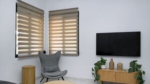 A modern living room with a grey armchair, a round wooden table, a TV mounted on the wall, and a wooden cabinet with plants. - El Dorado Corner (Chania)