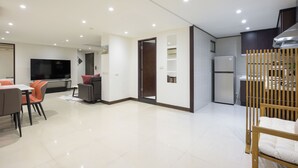 A modern living room with a flat-screen TV, dining area with chairs, and a kitchen with a refrigerator and cabinets.