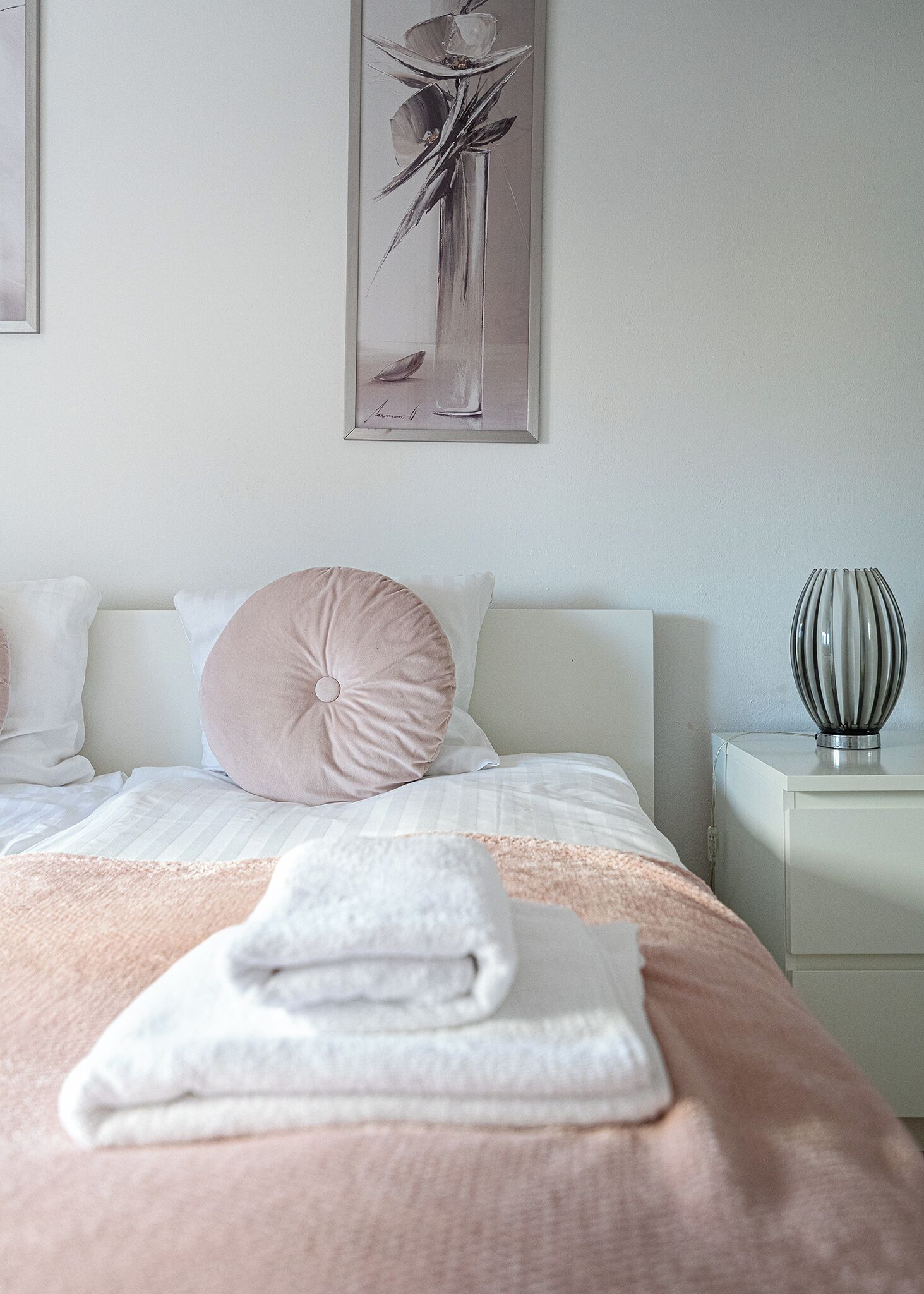 A neatly made bed with a pink pillow, a white pillow, and a folded white towel. There is a framed artwork above the bed and a metallic vase on a bedside table.