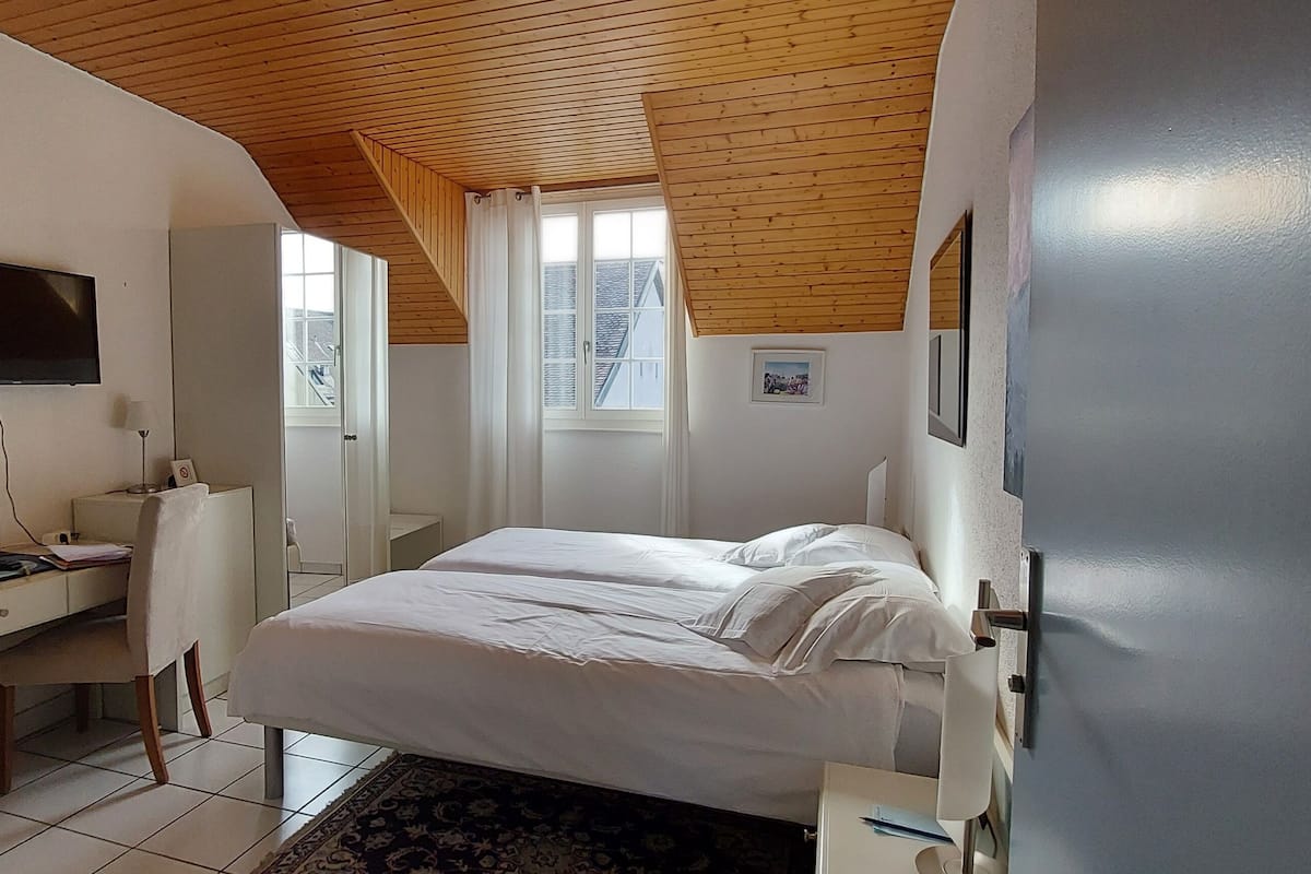 Quarto com cama, escrivaninha e cadeira. Tem uma janela com cortinas e um quadro na parede.