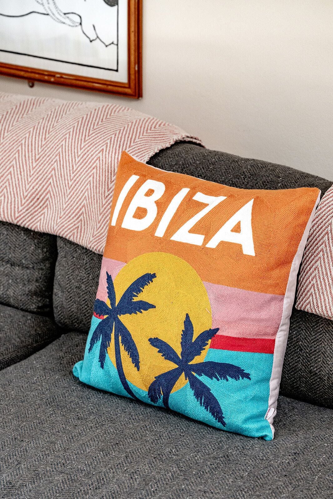 A cushion with 'IBIZA' text and a tropical design, placed on a grey sofa with patterned cushions.