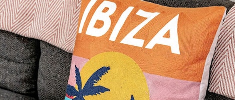 A cushion with 'IBIZA' text and a tropical design, placed on a grey sofa with patterned cushions.