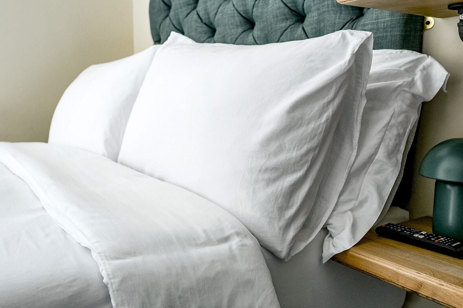 Una cama bien hecha con sábanas blancas, un cabecero tapizado en verde y una mesita de noche con un control remoto.