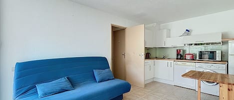 A compact living space with a blue sofa, a small wooden table, and a kitchenette with basic appliances.