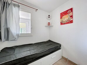 A small room with a bed, a window with curtains, a shelf with a can, and a painting on the wall. - Seafront apartment with terrace and pool (Saint-Cyprien)