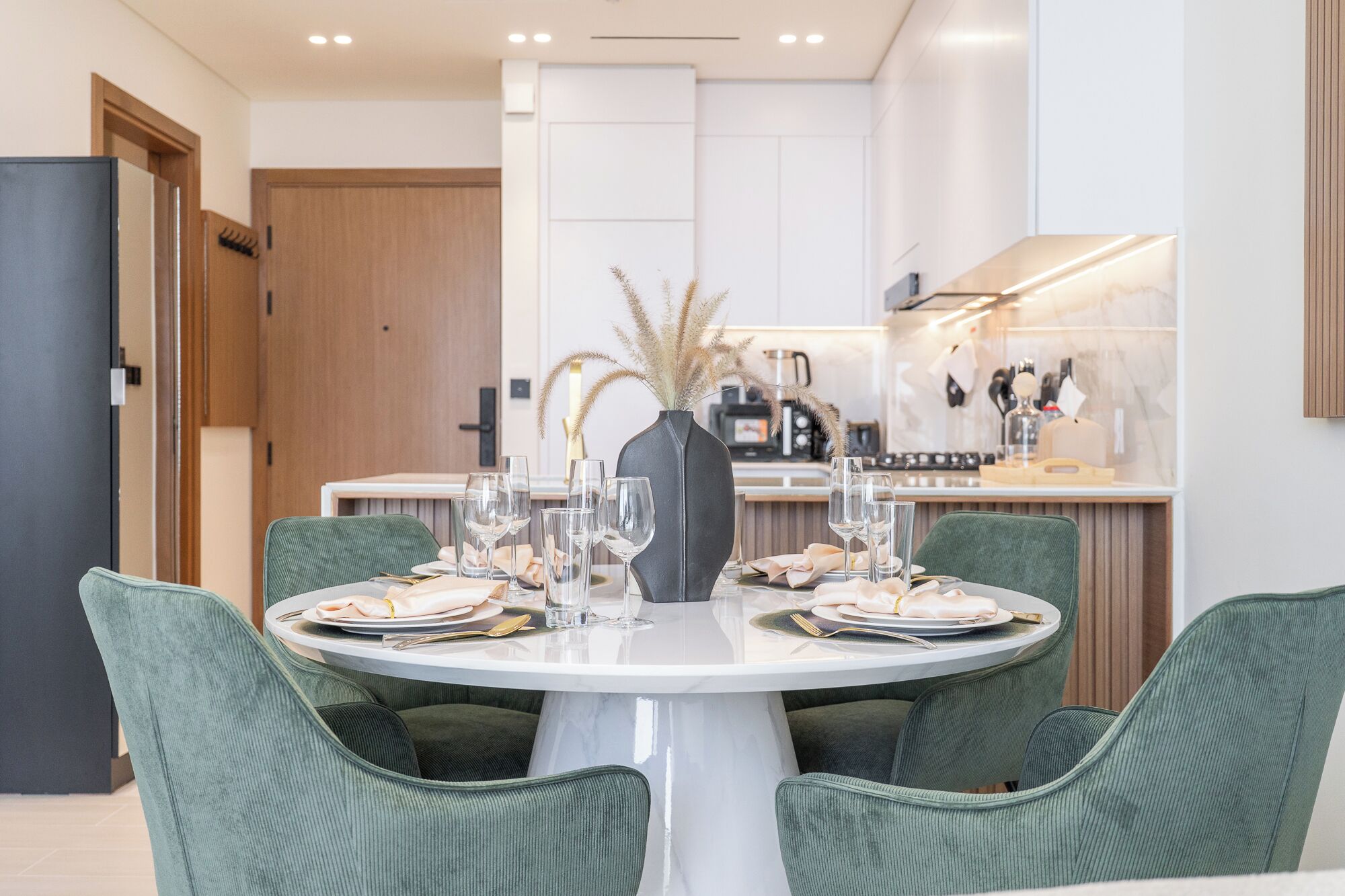 Un coin repas avec une table ronde dressée pour quatre personnes, entourée de chaises recouvertes d’un tissu vert. Un vase contenant des herbes de la pampa séchées est placé au centre.