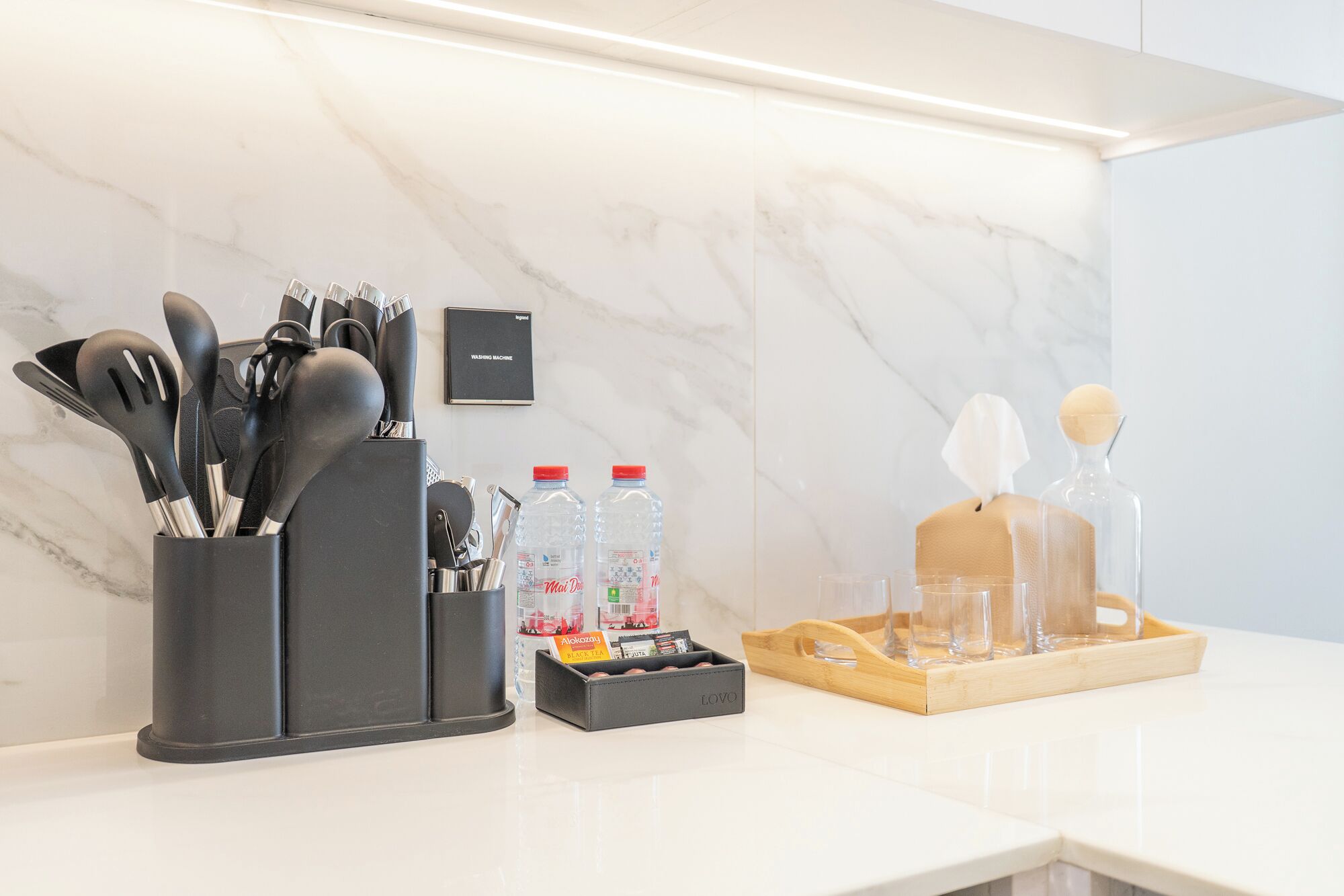 En køkkenbordplade med redskaber, en vandflaske, en sæbedispenser og en bakke med glas.