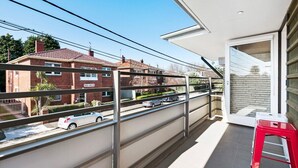 A balcony with a metal railing, a red stool, and a view of a residential street with cars and houses.