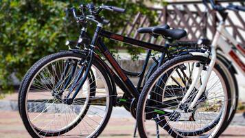 Una bicicleta estática con estructura negra y asiento, estacionada sobre superficie pavimentada, con cerca de madera y vegetación al fondo.