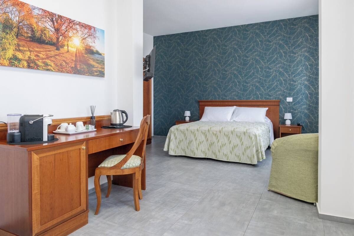 Une chambre d’hôtel avec un bureau en bois, un lit recouvert d’une couvre-lit à motifs de feuilles vertes, une chaise et une table de chevet avec une lampe.