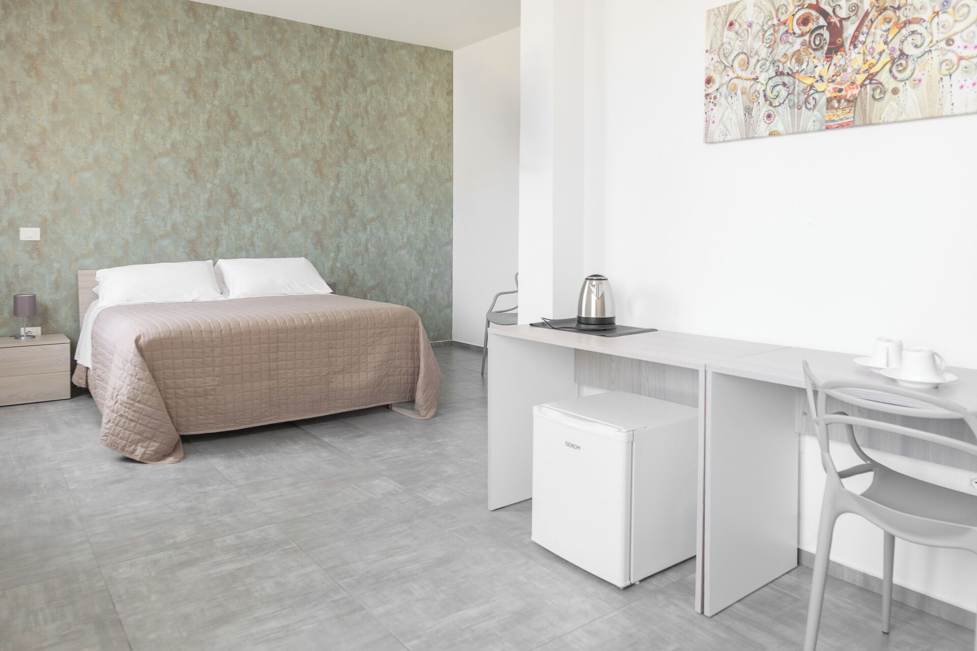 A modern hotel room with a bed, a desk, a chair, a small fridge, and a coffee maker.
