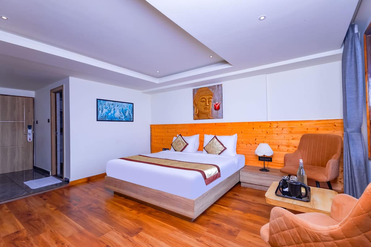 A modern hotel room with a wooden floor, a large bed with white bedding and decorative pillows, a wooden headboard, a small table with a bottle and a cup, and a chair.