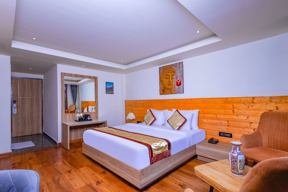 A hotel room with a wooden headboard, a bed with white bedding, a bedside table with a vase, a chair, and a desk with a mirror and a picture on the wall.