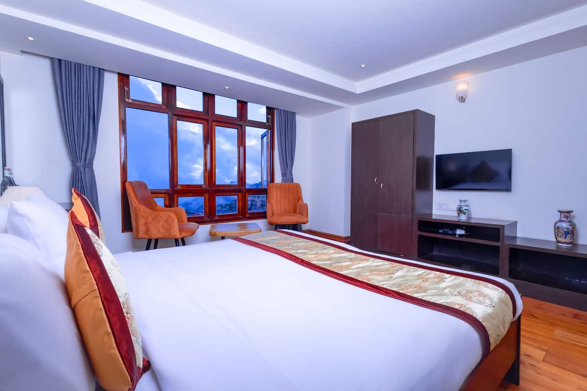A modern hotel room with a large bed, two chairs, a wardrobe, a TV, and a view of the sky through the window.