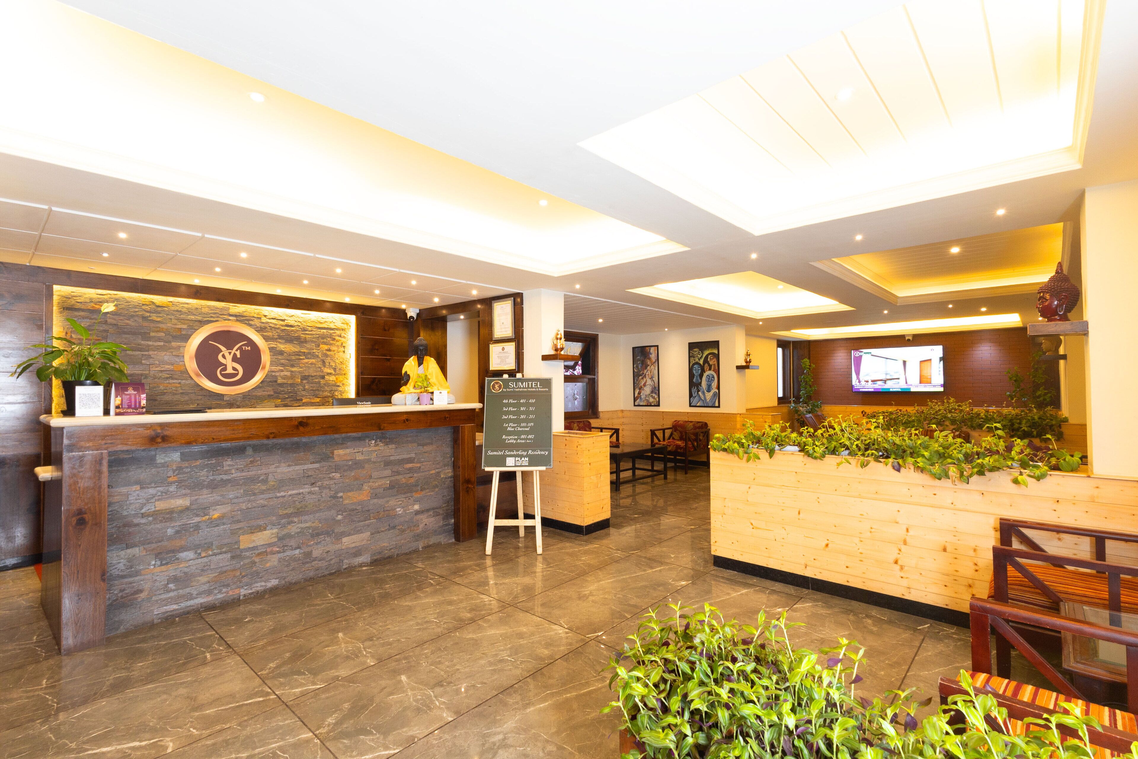 A hotel reception area with a stone reception desk, a wooden counter, and a seating area with plants.