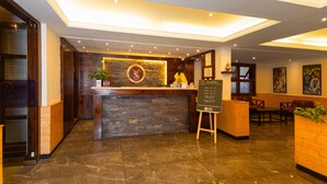 A reception area with a wooden counter, a stone wall, and a ceiling with recessed lighting. There are seating areas with wooden tables and chairs, and a signboard with information. - SUMITEL SANDERLING (Darjeeling)