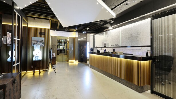 Zona de recepción de hotel con diseño moderno, que incluye un mostrador de recepción, un panel de pared de madera y una entrada con puerta de vidrio.