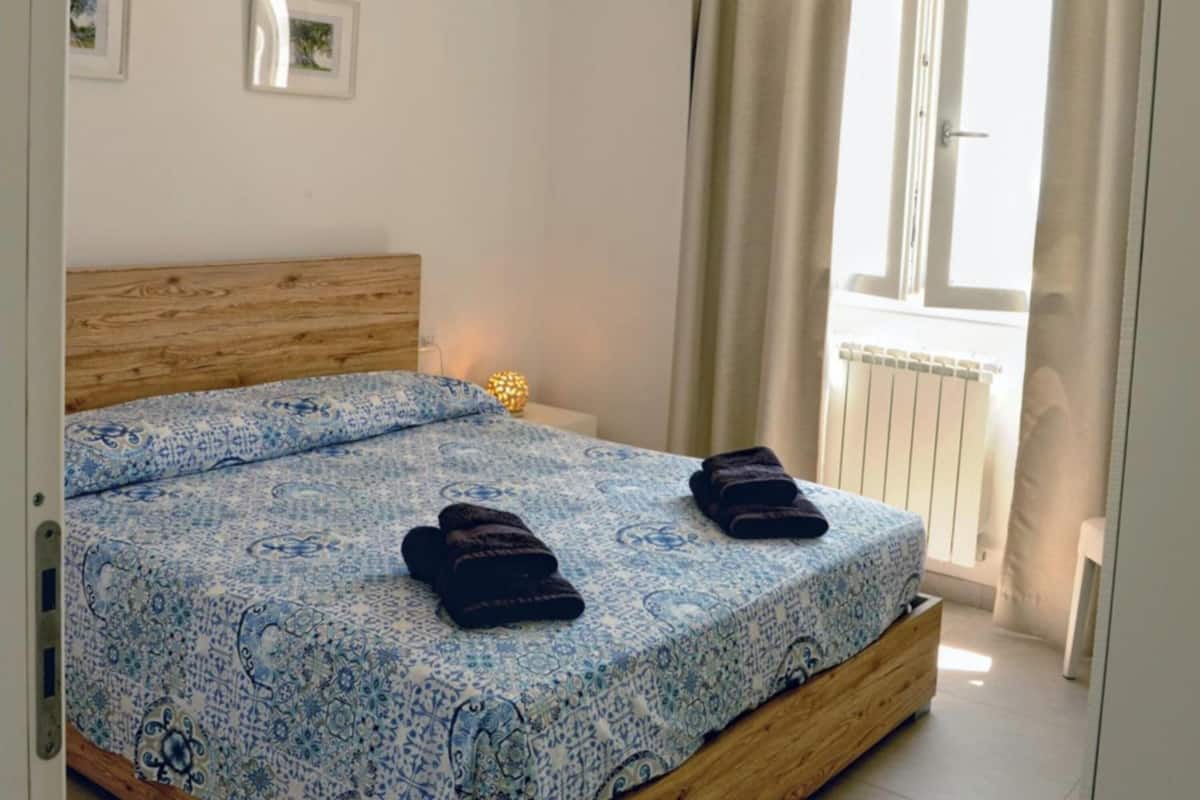 Quarto com cabeceira de madeira, colcha estampada e duas toalhas sobre a cama.